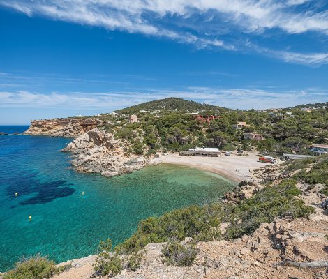 Panorama van Cala Carbó op Ibiza — kleine beschutte baai met turquoise water en pijnbomen langs de rotsige rand
