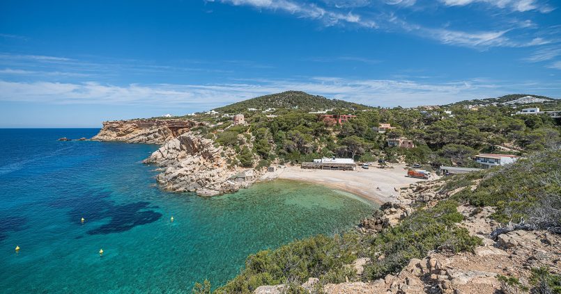 Panorama van Cala Carbó op Ibiza — kleine beschutte baai met turquoise water en pijnbomen langs de rotsige rand
