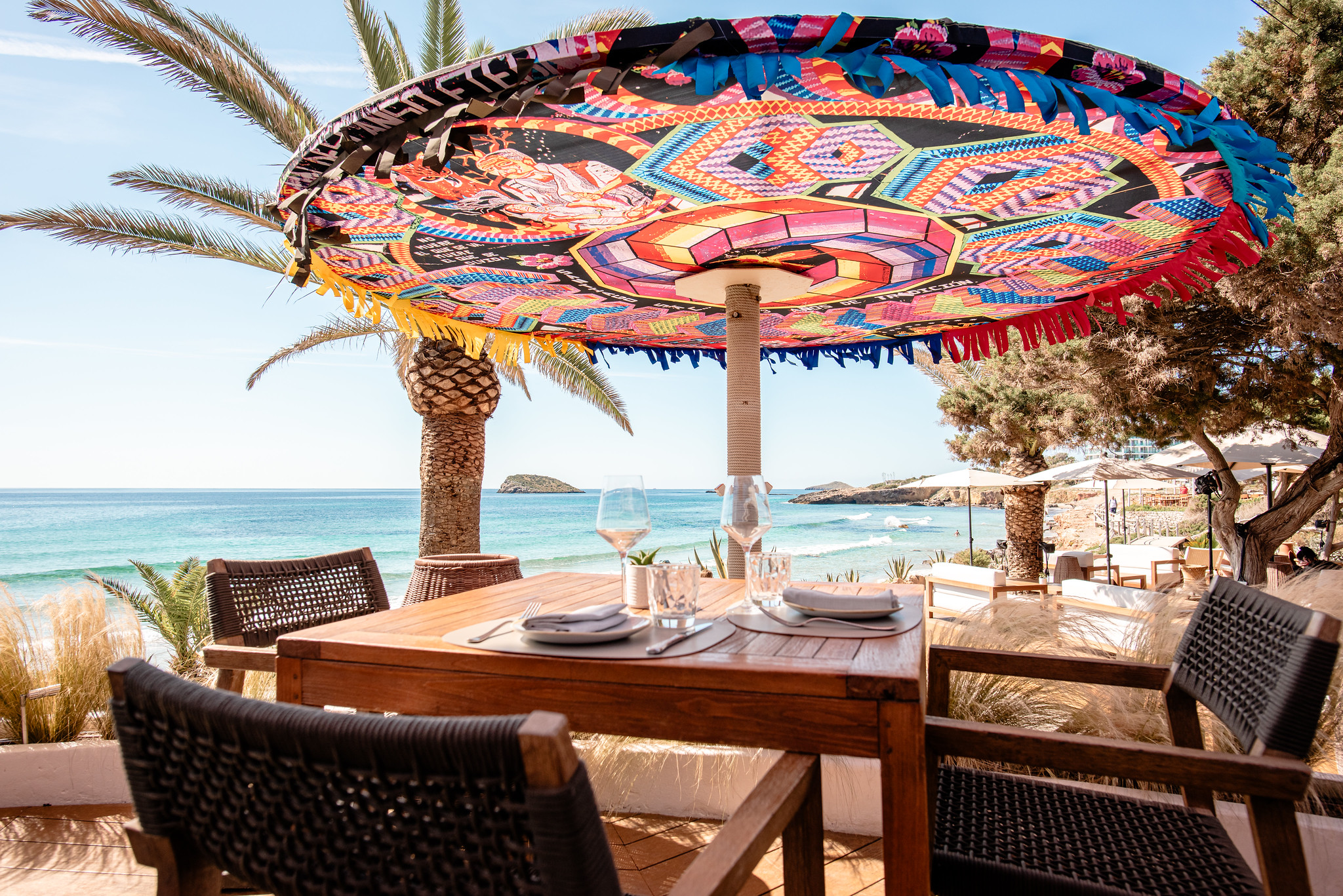 Aiyanna Ibiza: strandrestaurant met Levantijnse keuken aan Cala Nova 2 Een gedekte tafel onder een kleurrijke handgemaakte Guatemalteekse parasol bij Aiyanna, met uitzicht op de baai van Cala Nova, Ibiza