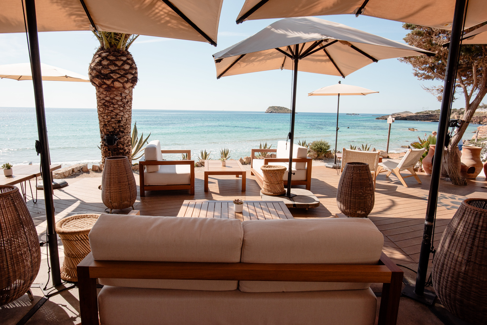 Aiyanna Ibiza: strandrestaurant met Levantijnse keuken aan Cala Nova 1 De oceaanlounge van Aiyanna aan Cala Nova op Ibiza, met cremekleurige banken, parasols en uitzicht over de turquoise baai