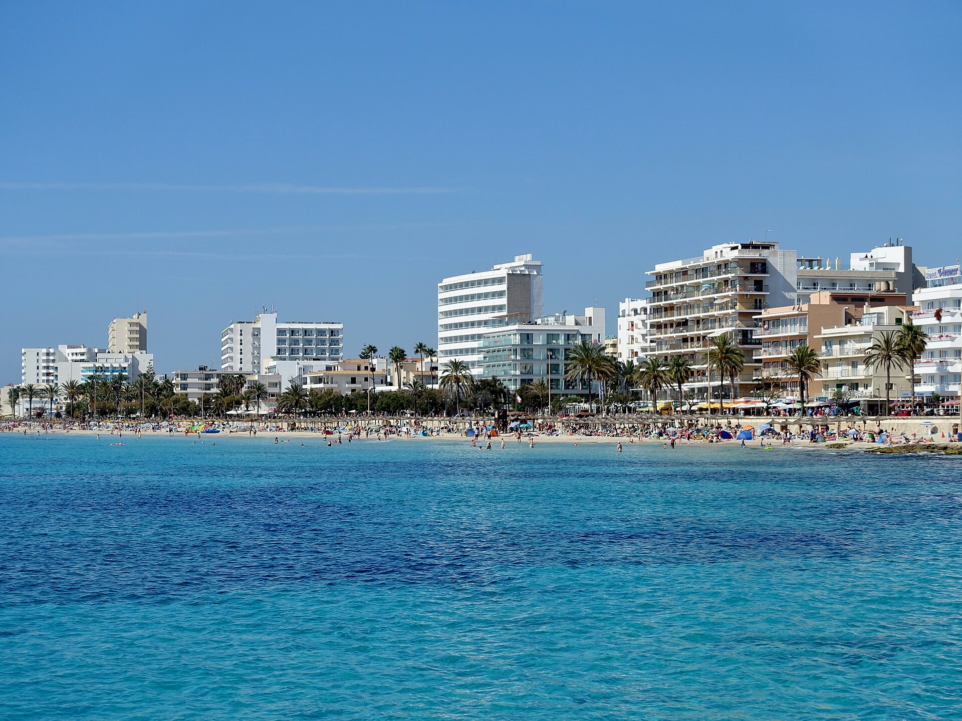 Het lange zandstrand van Cala Millor met turquoise water, strohutparasols, palmpromenade en hotels, oostkust van Mallorca