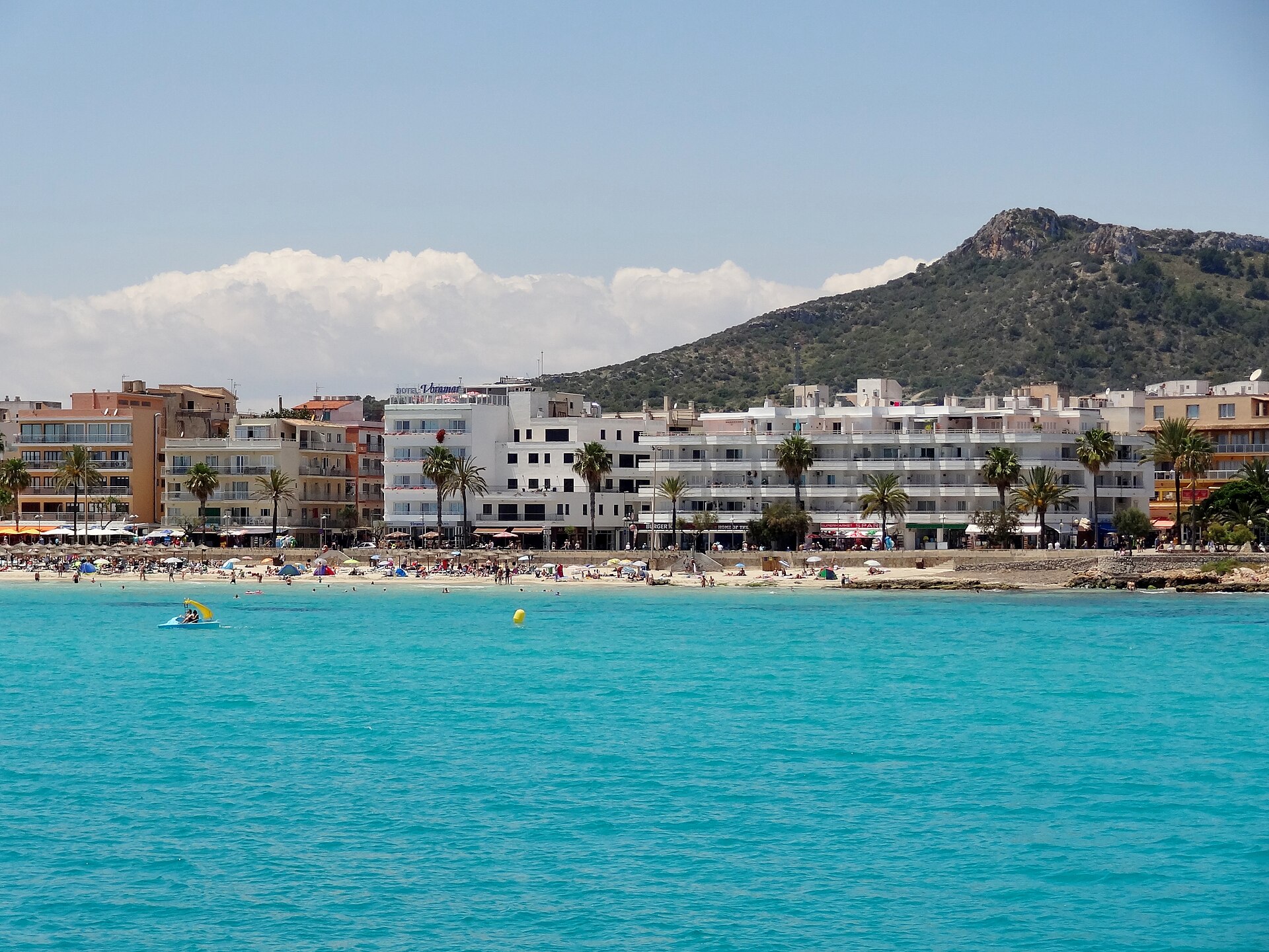 Helder turquoise water bij Cala Millor met een tretbootje, het strand met parasols en een groene berg op de achtergrond, Mallorca