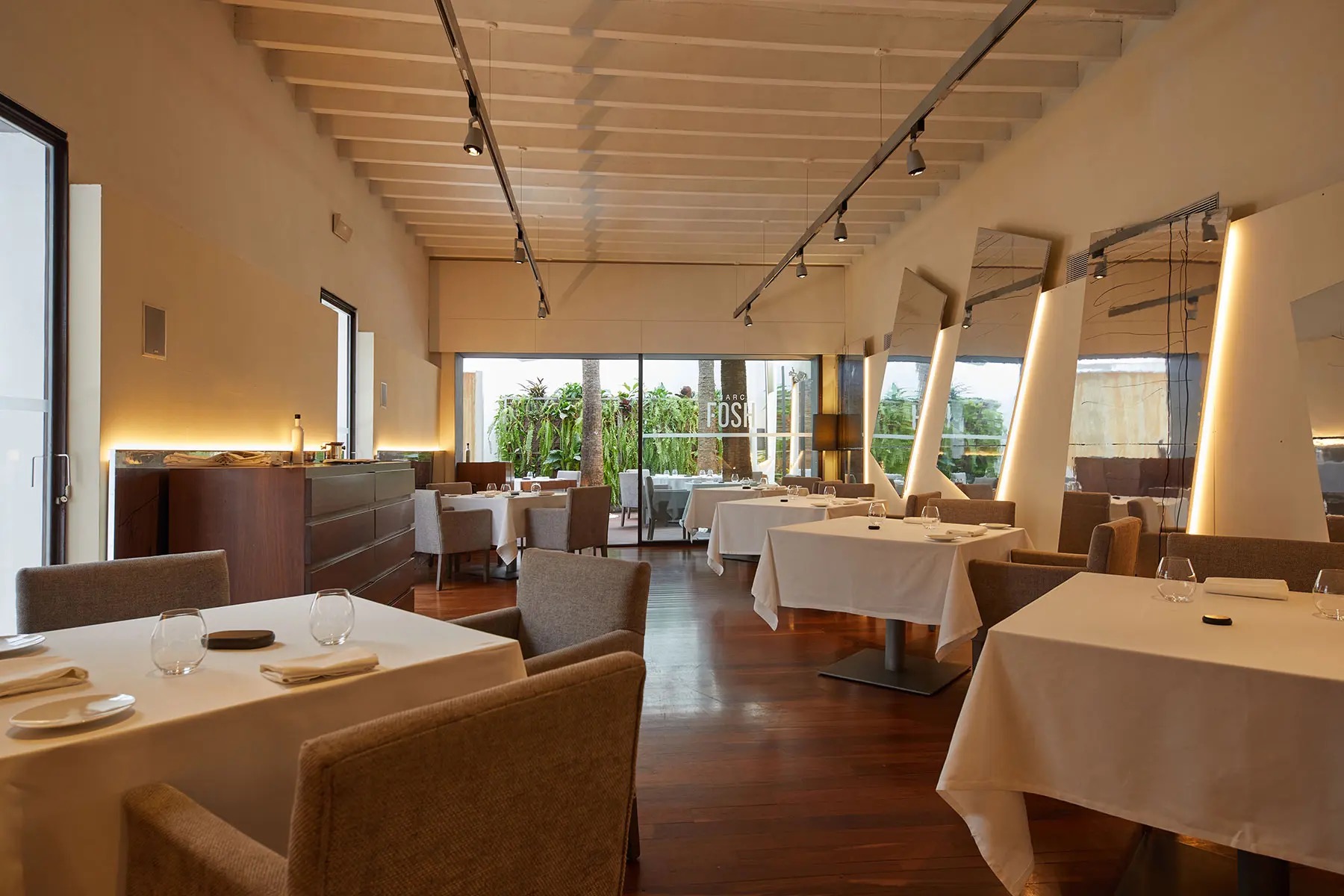 De elegante, warm verlichte eetzaal van Marc Fosh met witte tafels en doorkijk naar het groene binnenplaatsterras, Palma