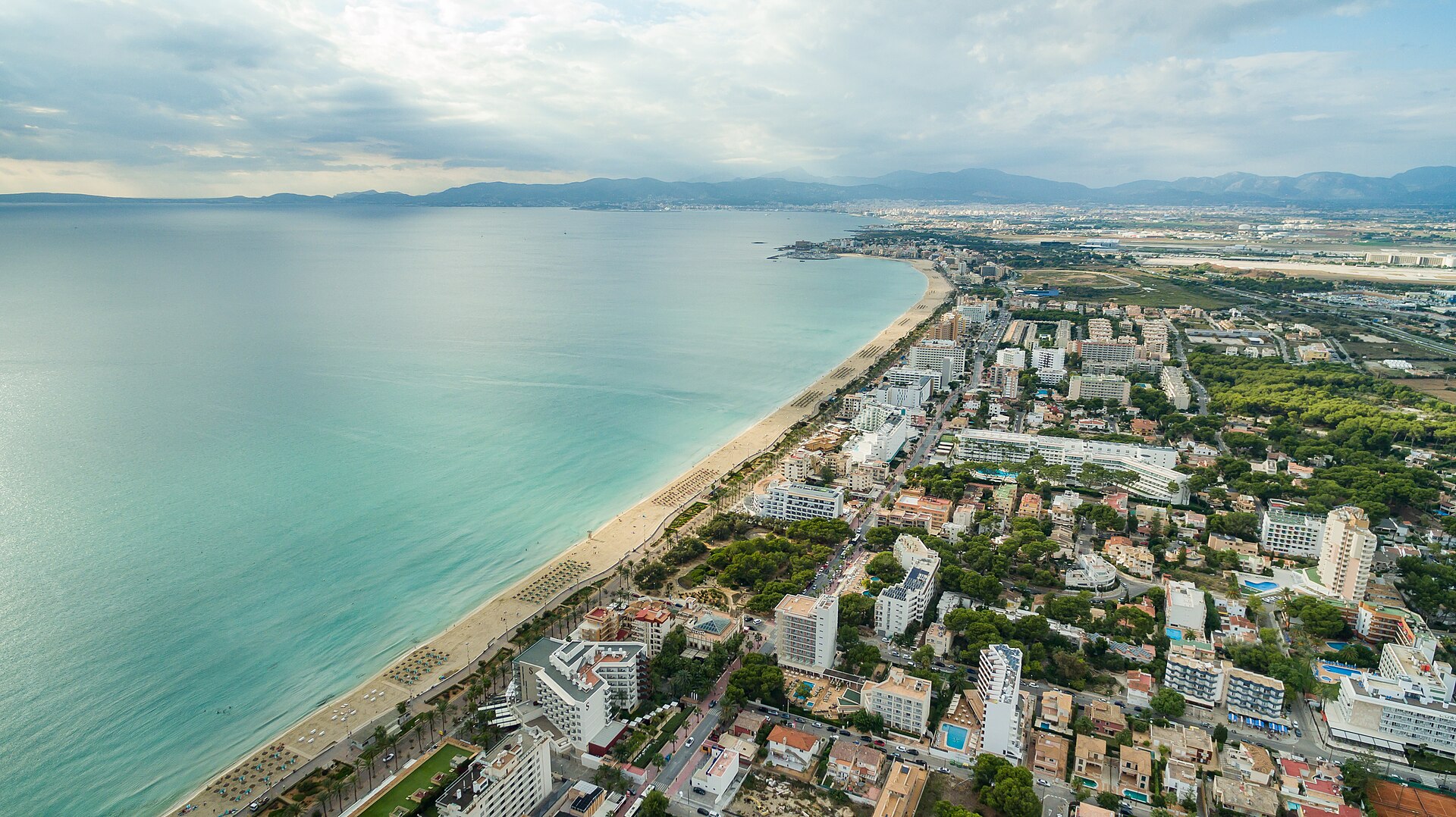 Luchtfoto van het kilometerslange goudkleurige strand van Playa de Palma met promenade, parasols en de Serra de Tramuntana in de verte, Mallorca
