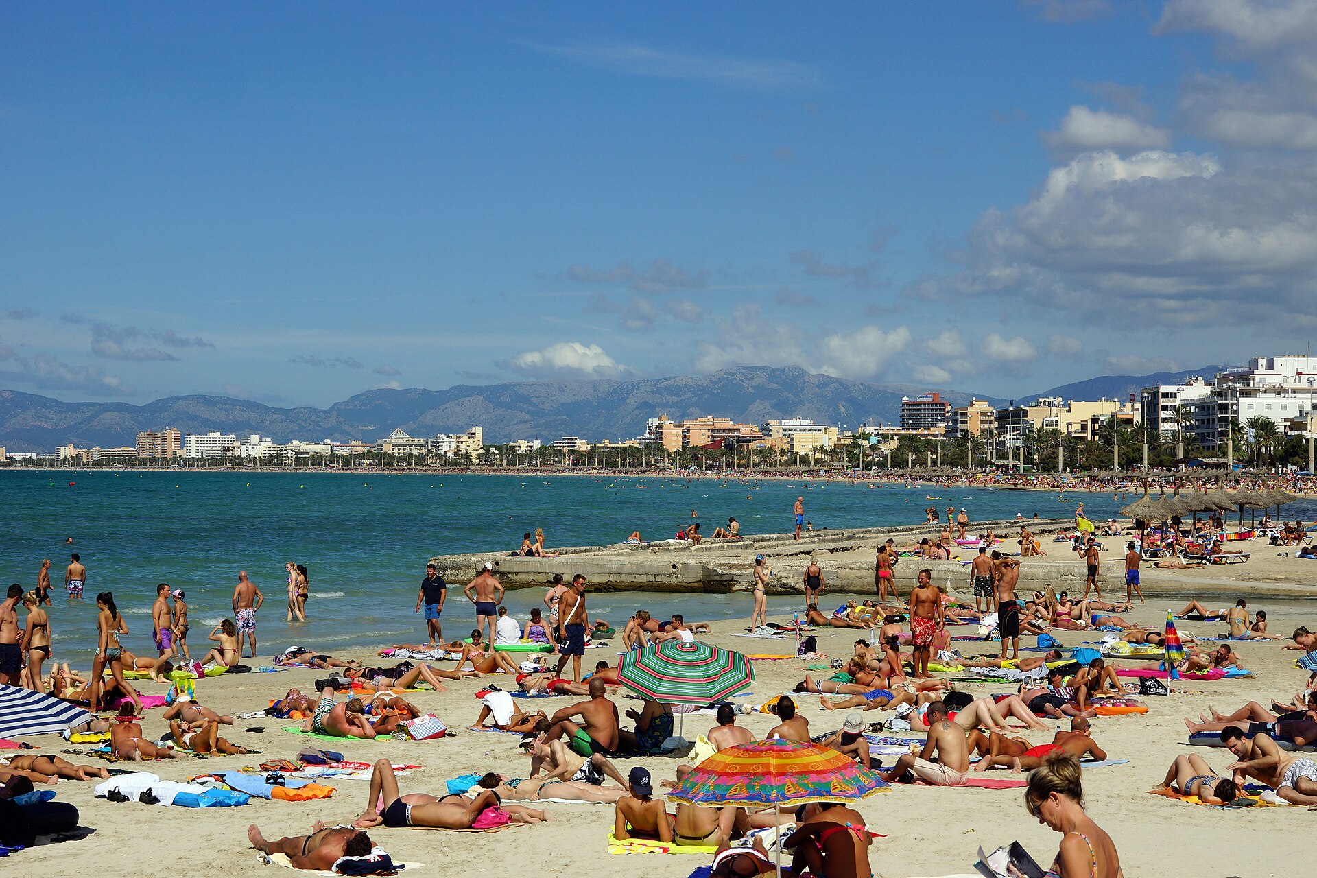 Druk zomerstrand van Playa de Palma met veel zonnende badgasten, kleurrijke parasols en de promenade en bergen op de achtergrond, Mallorca