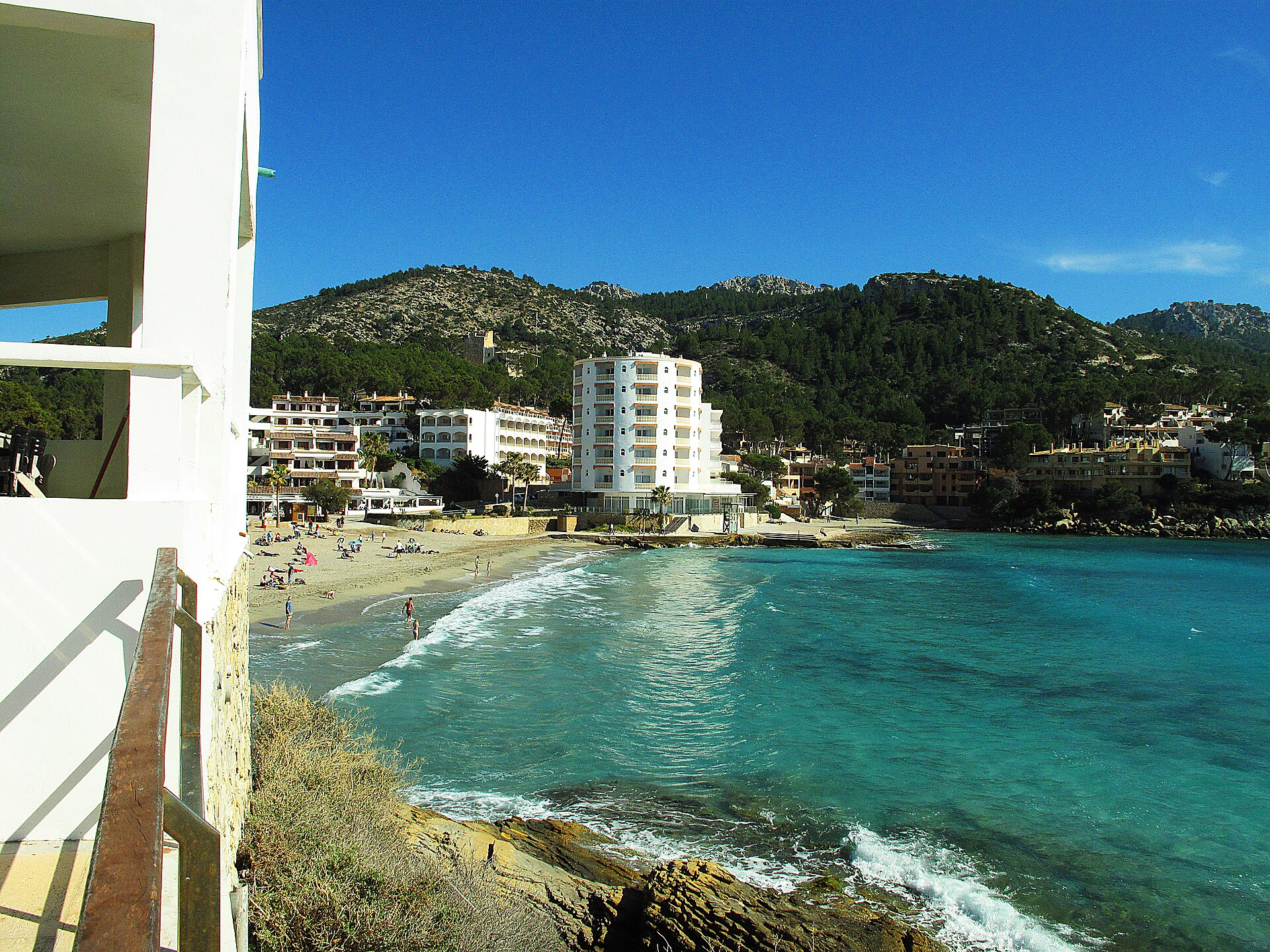 Het zandstrand van Sant Elm met turquoise water, badgasten en de dorpsbebouwing tegen de beboste heuvels, Mallorca