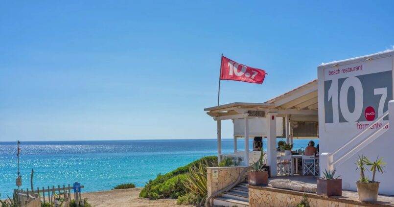 De witte strandlounge 10.7 met de vlag boven Platja de Migjorn en de turquoise zee
