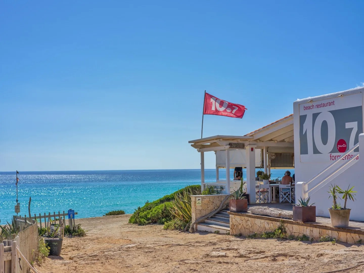 10.7 Formentera: zonsondergang en fusionkeuken aan Platja de Migjorn 1 De witte strandlounge 10.7 met de vlag boven Platja de Migjorn en de turquoise zee