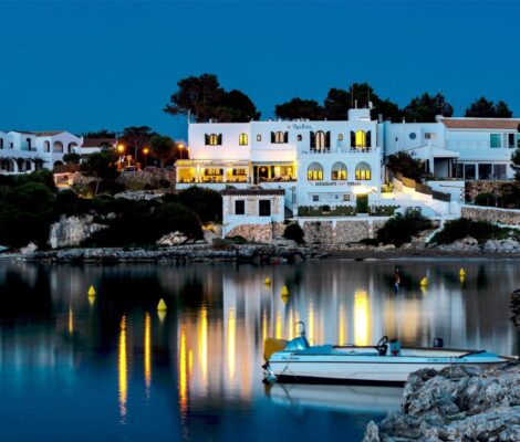 Het verlichte Menorcaanse huis van Bahía Santandría weerspiegeld in de cala van Santandría bij schemering