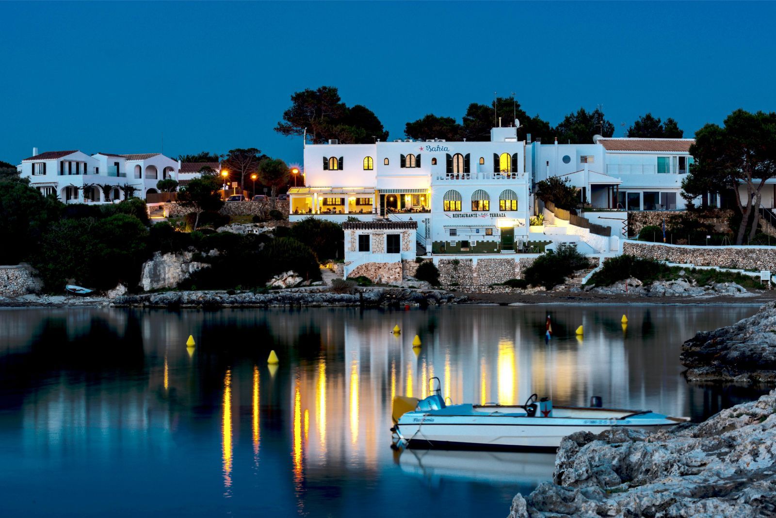 Het verlichte Menorcaanse huis van Bahía Santandría weerspiegeld in de cala van Santandría bij schemering