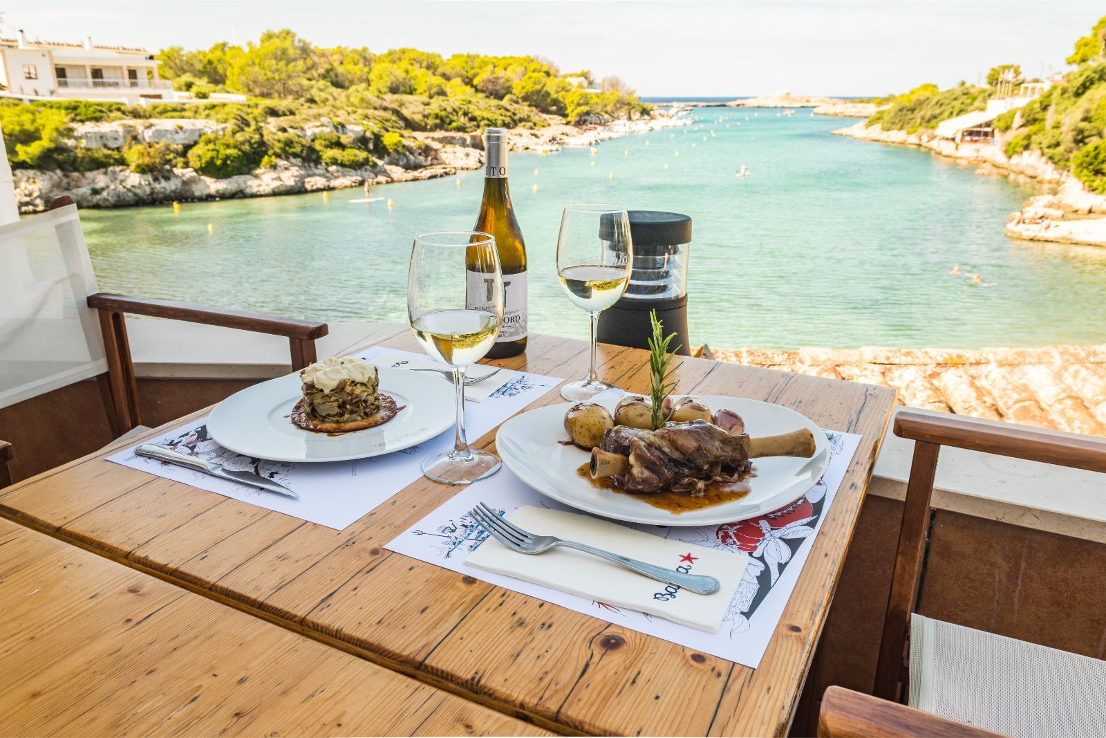 Een gedekte tafel op het terras van Bahía Santandría met gerechten en wijn en uitzicht over de cala