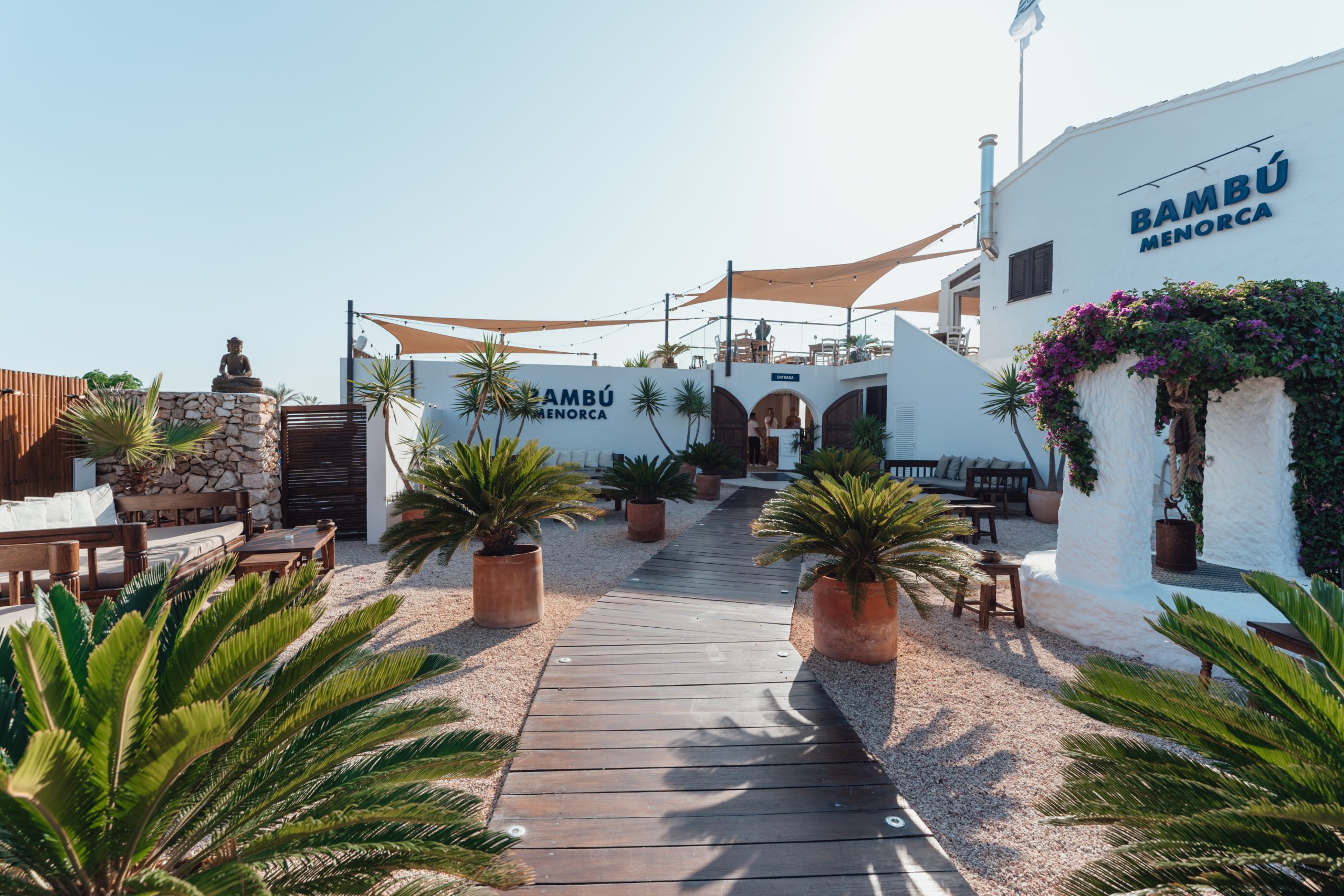 De witte ingang en het houten terras van Bambú Menorca met palmen