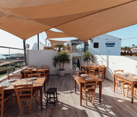 Het terras met gedekte tafels van Bambú Menorca onder zonnezeilen met de zee erachter