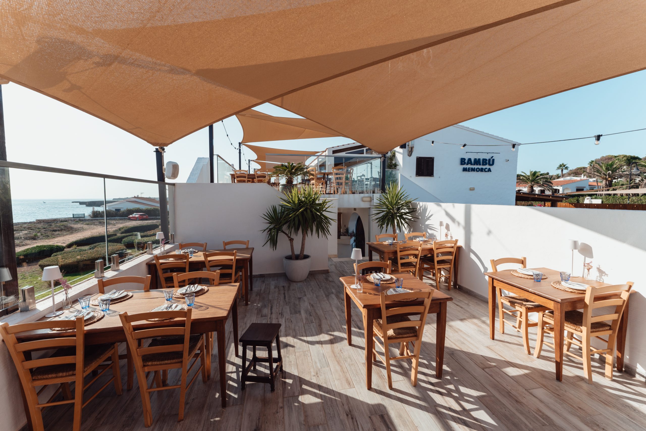 Het terras met gedekte tafels van Bambú Menorca onder zonnezeilen met de zee erachter
