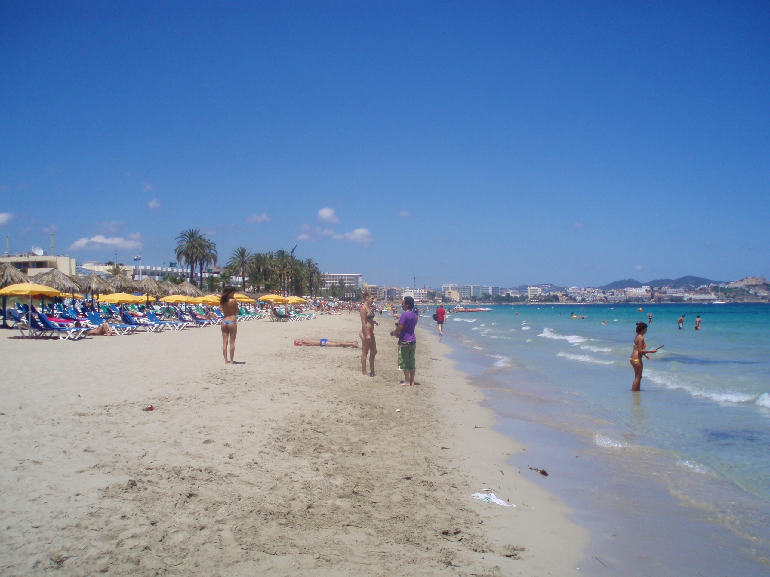 Beachclubs op Ibiza: waar ze zitten, seizoen en reserveren 2 Het strand van Playa d'en Bossa op Ibiza met links de gebouwen van beachclub Bora Bora en badgasten in het ondiepe water