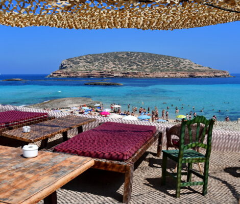 Terras van een strandbar boven Cala Comte op Ibiza met lage tafels, kussens en uitzicht op het turquoise water en de eilandjes