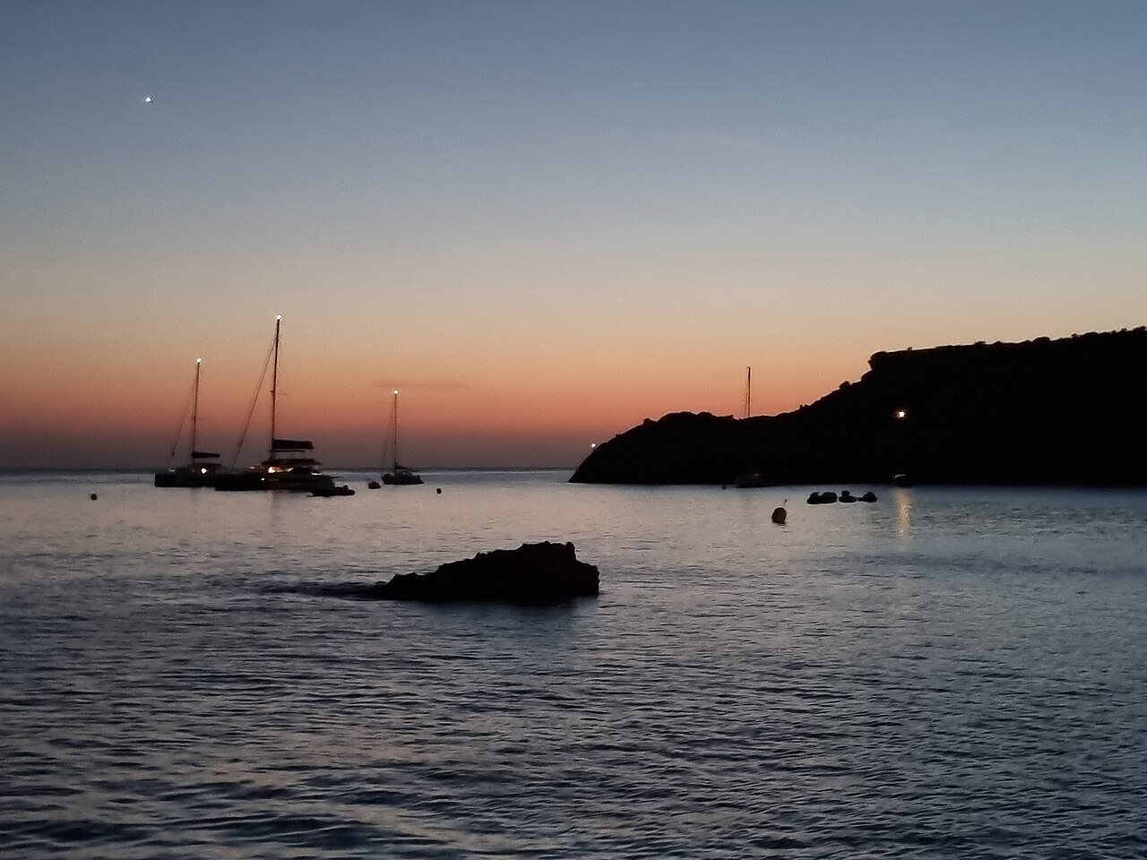 Beachclubs op Ibiza: waar ze zitten, seizoen en reserveren 5 Zonsondergang boven Cala Tarida op Ibiza met een zeilboot in silhouet op het stille water