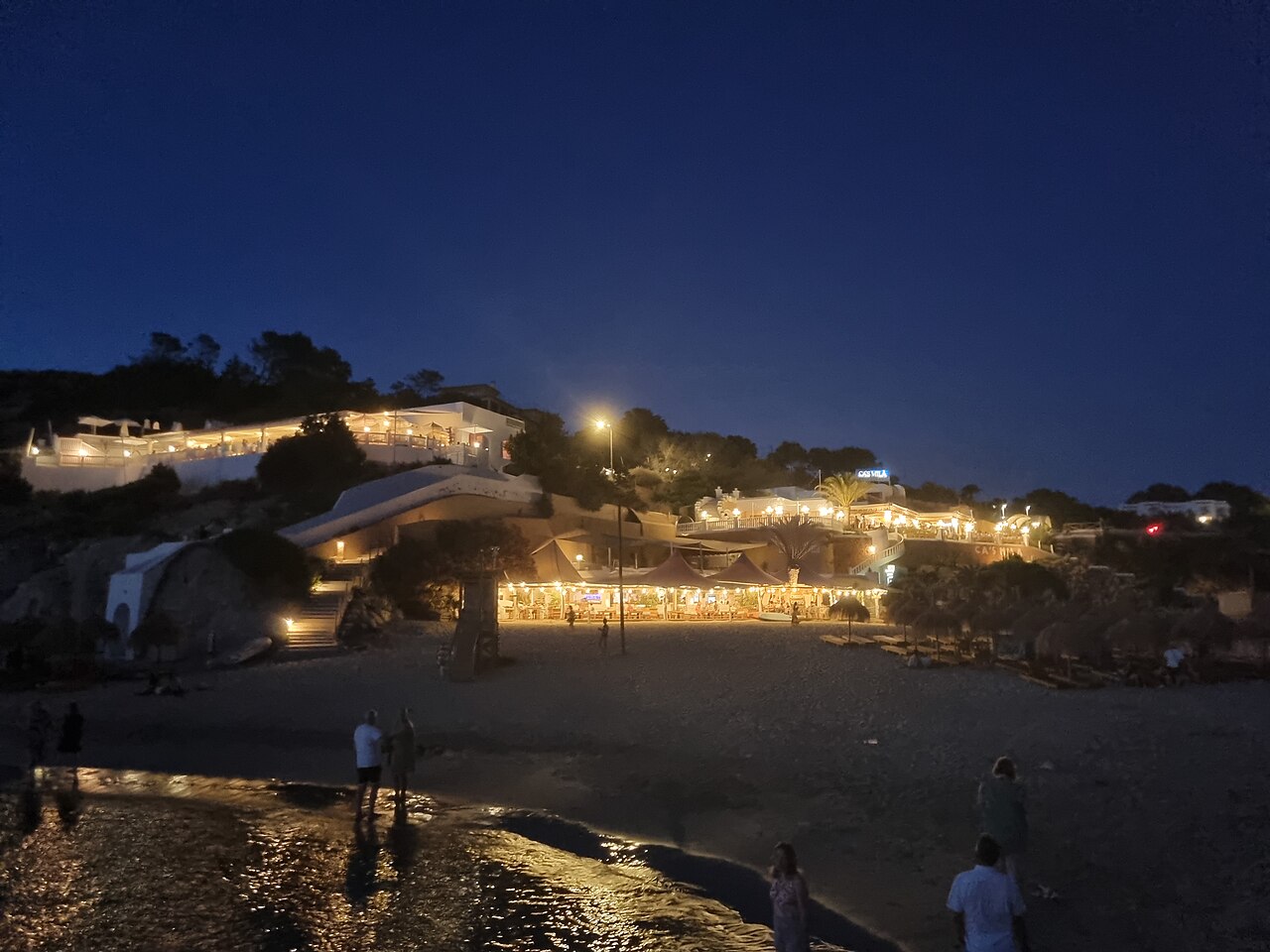 Beachclubs op Ibiza: waar ze zitten, seizoen en reserveren 6 De verlichte gebouwen van Cala Tarida op Ibiza in de avond, weerspiegeld in een rotspoel aan de waterkant