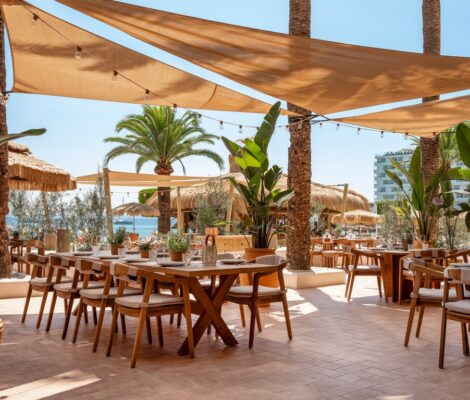 Het terras van Beso Beach Mallorca met houten tafels onder zonnedoeken en palmen aan het strand van Palmanova