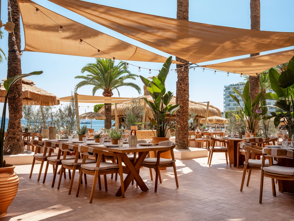 Beso Beach Palmanova (Mallorca): barefoot-luxury aan het strand 1 Het terras van Beso Beach Mallorca met houten tafels onder zonnedoeken en palmen aan het strand van Palmanova