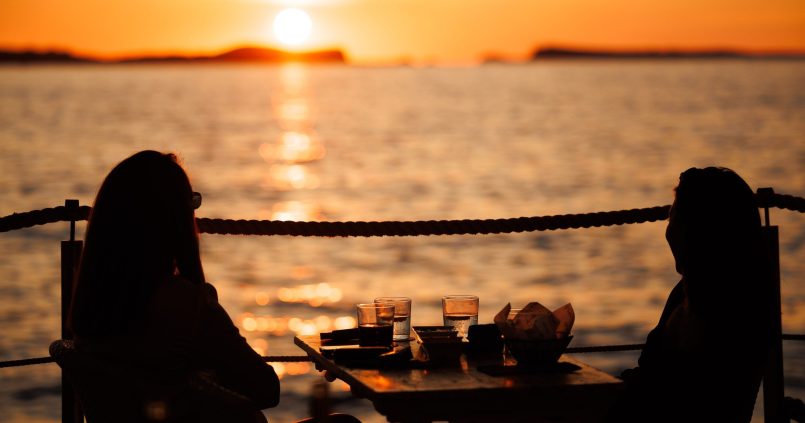Twee gasten als silhouet aan een tafel op het terras van Café del Mar, met de zon die boven zee ondergaat