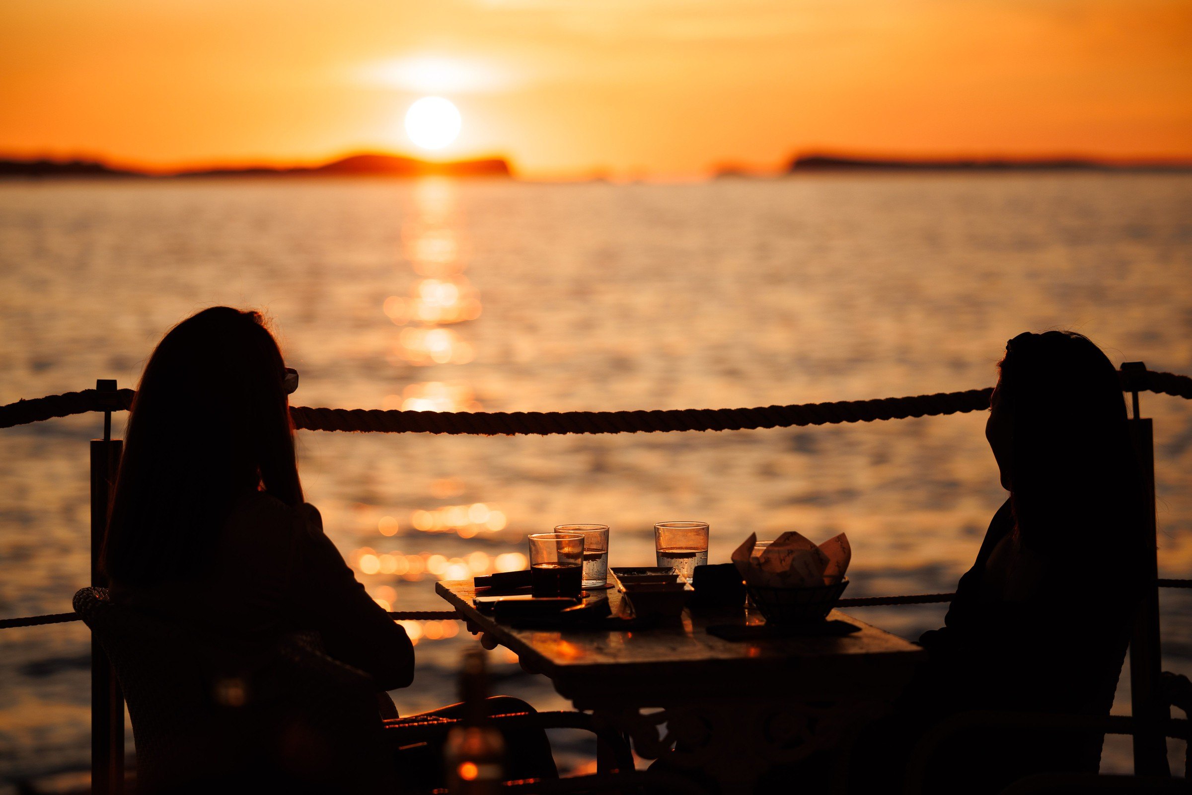 Twee gasten als silhouet aan een tafel op het terras van Café del Mar, met de zon die boven zee ondergaat