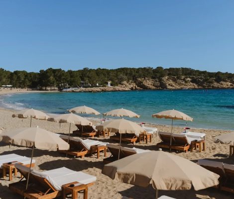 Het strand van Cala Bassa op Ibiza met de rij crèmekleurige parasols en houten ligbedden op het witte zand, turquoise water en de beboste kaap erachter