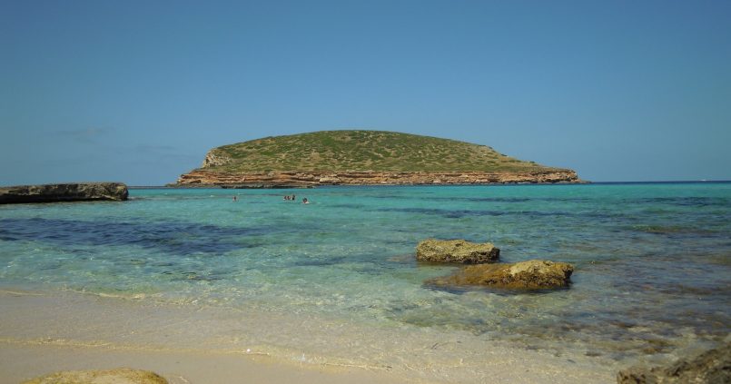 Het turquoise water van Cala Comte op Ibiza met het groene eilandje Illa des Bosc ervoor en zandbodem en rotsen op de voorgrond