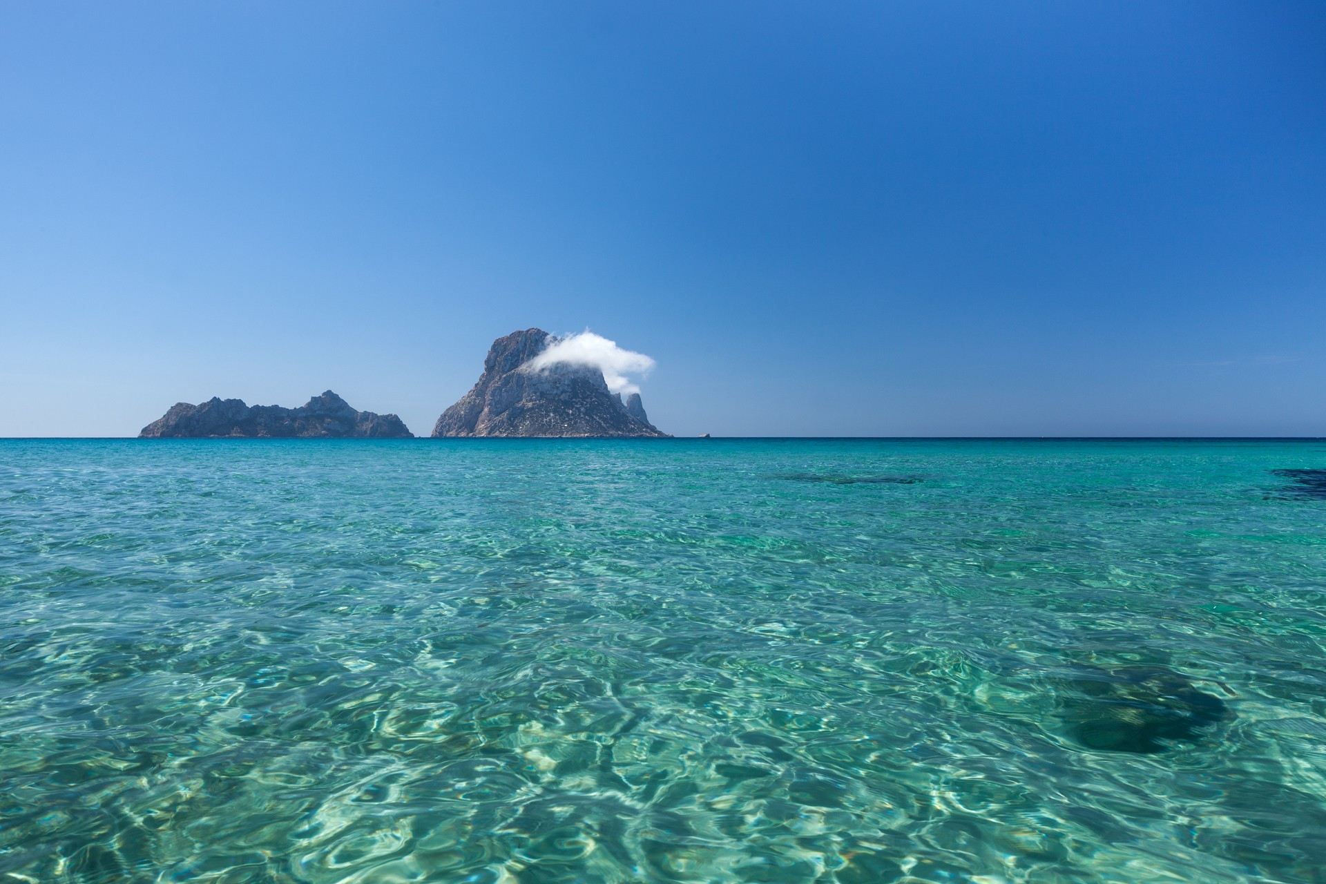 Helder turquoise water voor Cala d'Hort met de rotseilanden Es Vedranell en Es Vedrà aan de horizon