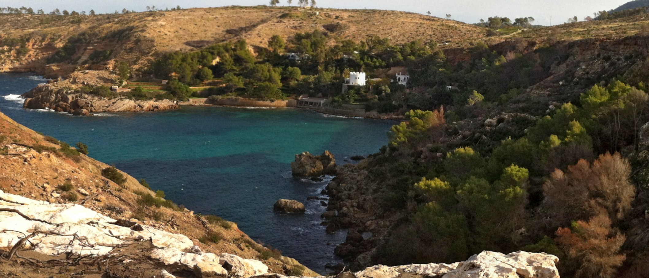 Cala es Canaret Ibiza: verborgen baai bij Portinatx, met de wandeling en de toegang in 2026 2 De binnenste cala van Es Canaret met de boothuizen, de keermuur langs het water en de villa met toren tussen de pijnbomen