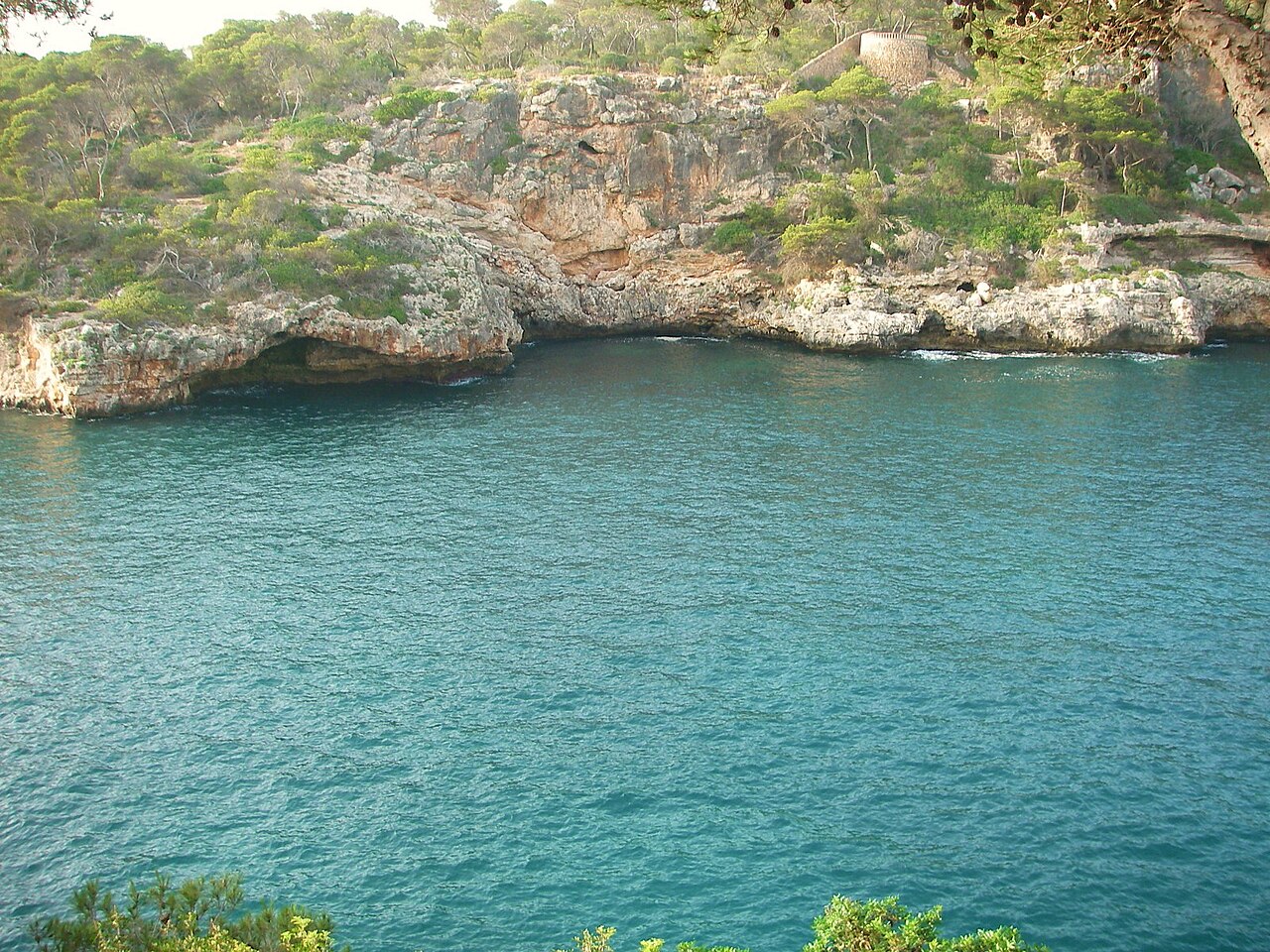 De rotskust bij de monding van Cala Figuera, met overhangende kalkkliffen boven het turquoise water en de ronde wachttoren Torre d'en Beu op de klif erboven