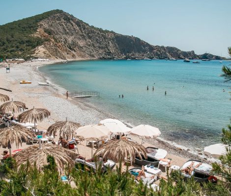 De baai van Cala Jondal met strohoedjes-parasols op het strand, turquoise water en de beboste landtong met rotswand erachter