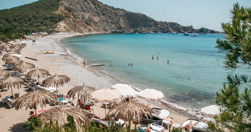 De baai van Cala Jondal met strohoedjes-parasols op het strand, turquoise water en de beboste landtong met rotswand erachter