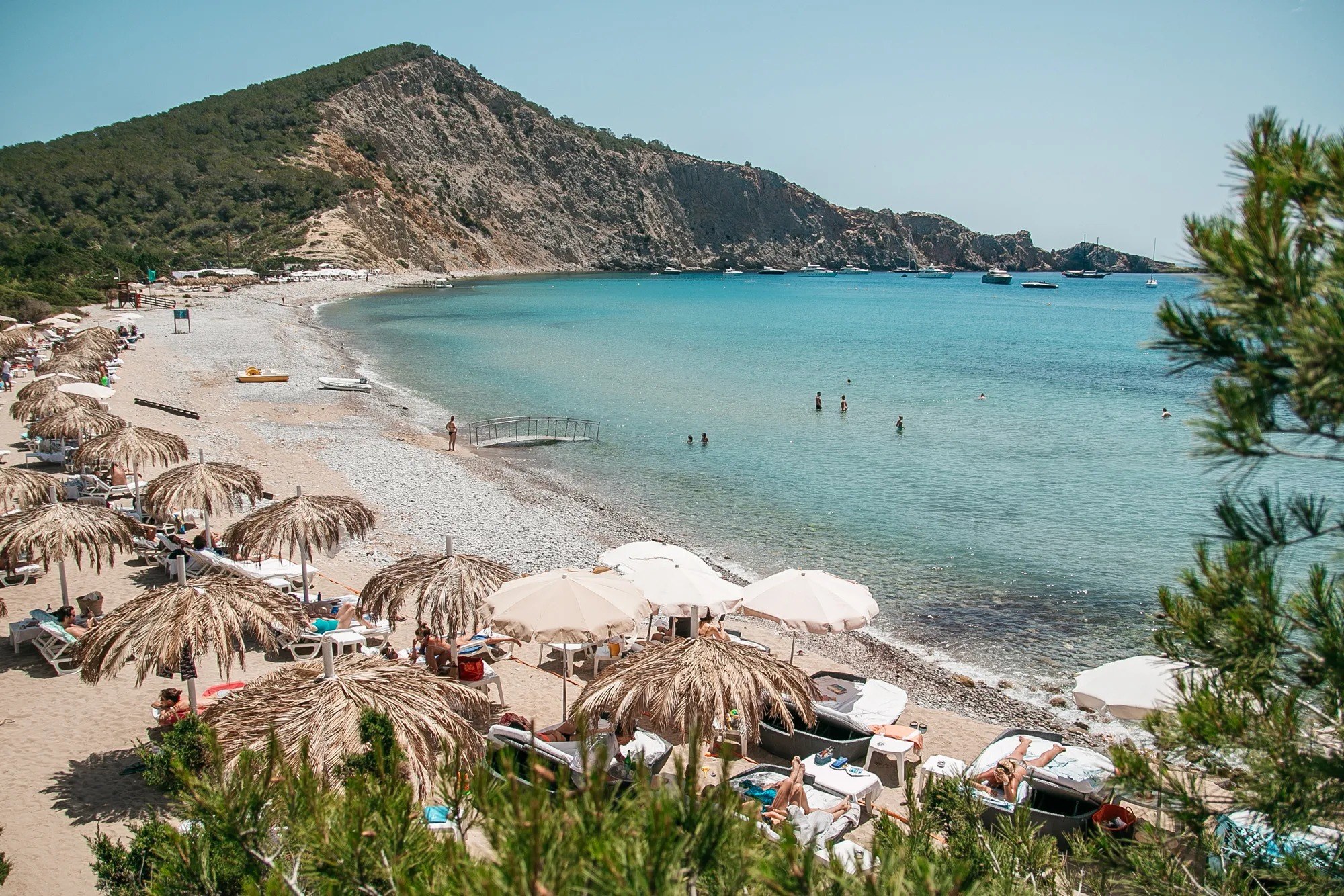 Cala Jondal Ibiza: het exclusieve jet-set-strand met Blue Marlin beach-club 1 De baai van Cala Jondal met strohoedjes-parasols op het strand, turquoise water en de beboste landtong met rotswand erachter