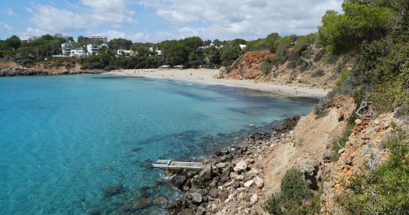 De baai van Cala Llenya op Ibiza met turquoise water, het zandstrand en roodbruine kliffen tussen de dennen