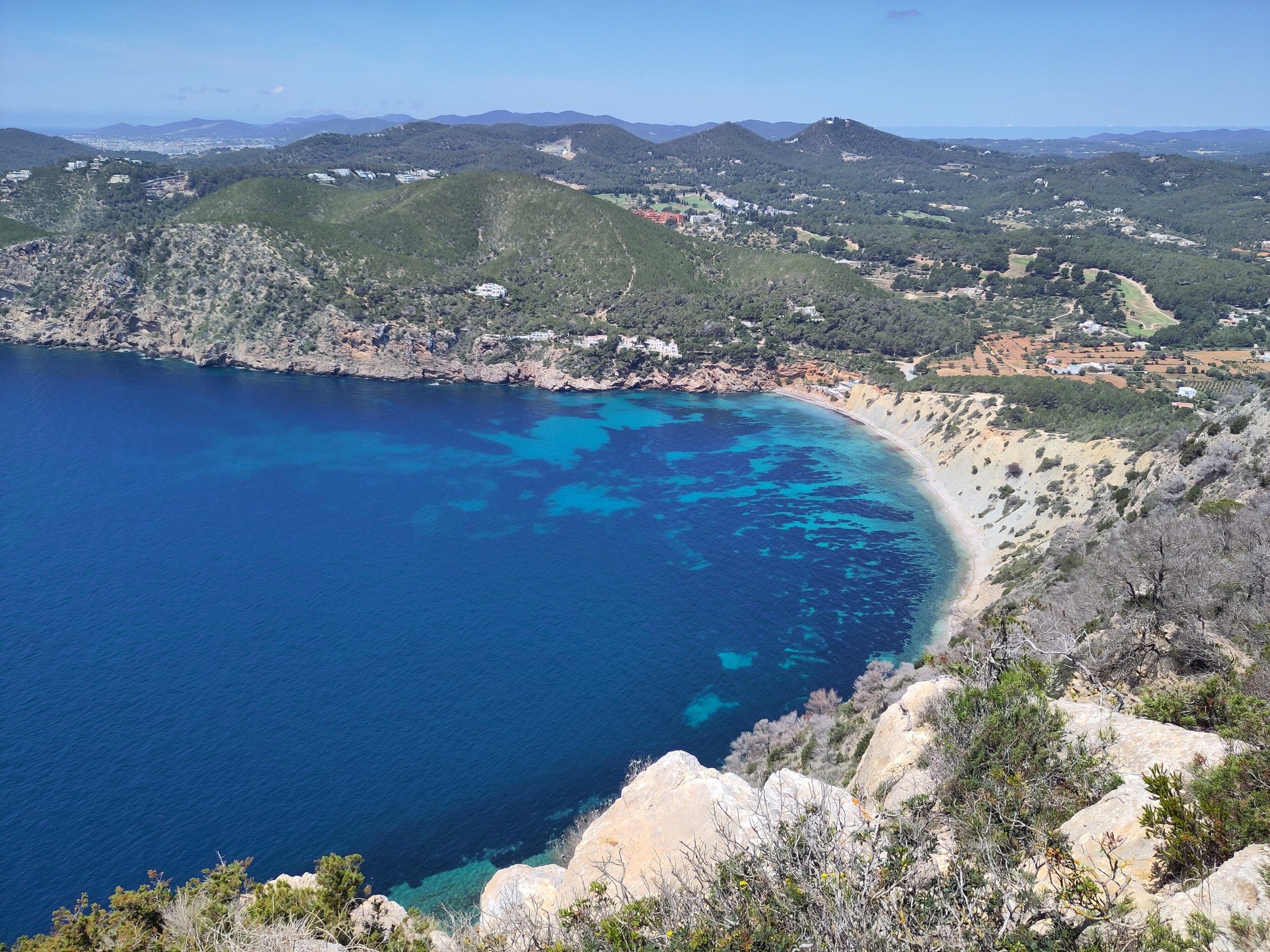 Uitzicht vanaf cap des Llibrell over diepblauw water en de baai van Sol d'en Serra, met de heuvels van Ibiza op de achtergrond