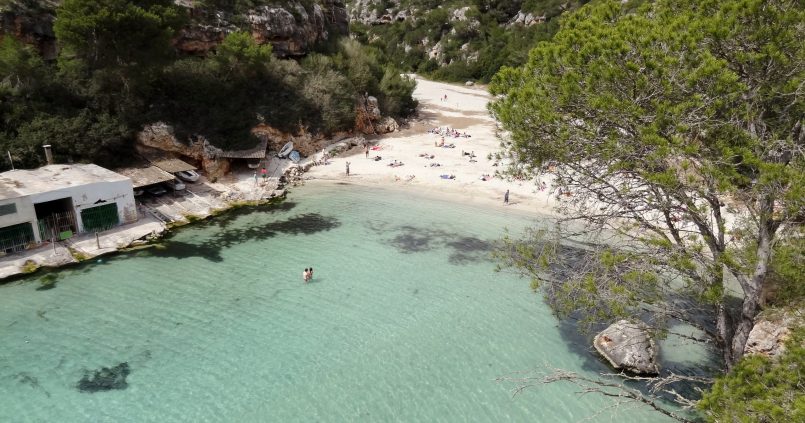 Het zandstrand van Cala Pi aan het eind van de smalle inham met helder turquoise water, boothuizen en beboste kliffen