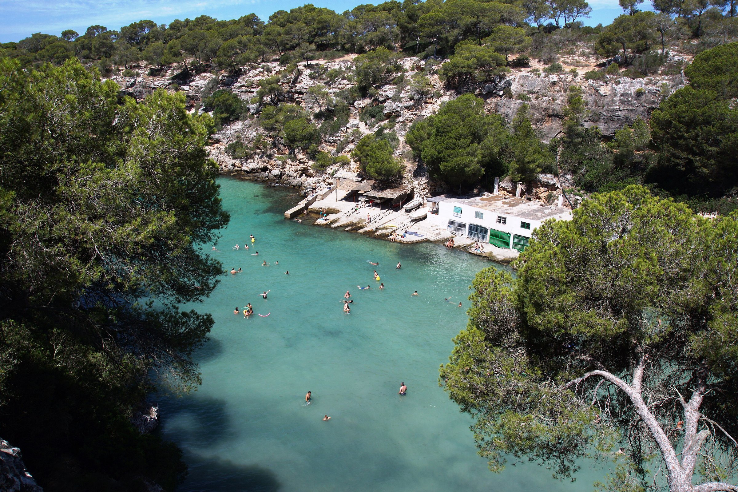 Zwemmers in het turquoise water van de smalle inham van Cala Pi met de witte boothuizen tegen de klif