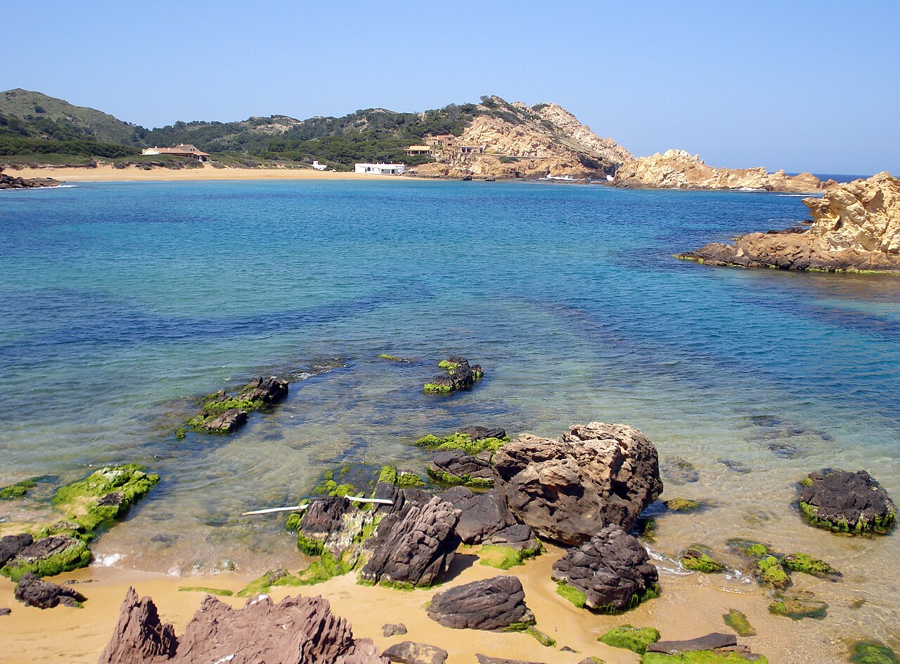 Het ondiepe water van Cala Pregonda op Menorca met rotsblokken en het roodgouden strand