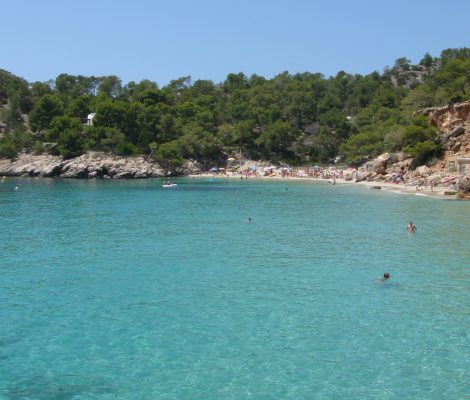 De baai van Cala Salada met turquoise water, pijnbos op de heuvels en vissershuisjes onder de rode rotswand