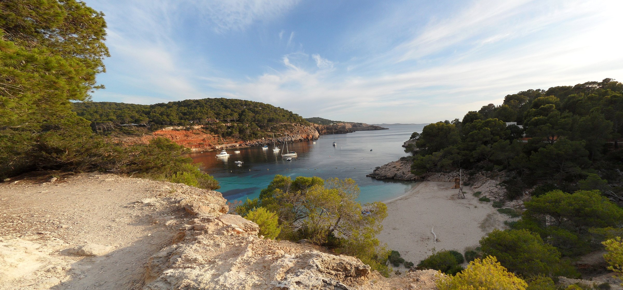 Cala Salada Ibiza: vissersbaai aan de rand van Es Amunts, met Cala Saladeta en de toegang in 2026 3 Het zandstrand van Cala Saladeta van bovenaf gezien, tussen pijnbomen, met rode kliffen aan de overkant van de baai
