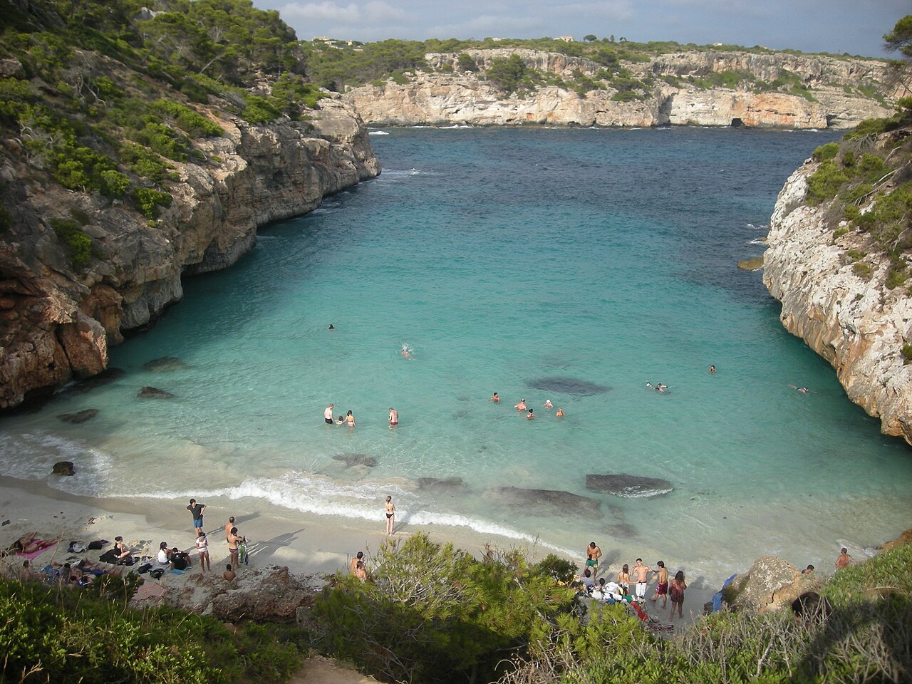 Het zandstrookje van Caló des Moro met badgasten, gezien vanaf het rotspad boven de baai