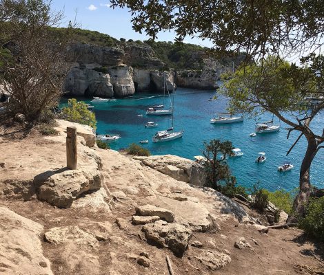 Een markeringspaal van het Camí de Cavalls op de rotsen boven Cala Macarella met boten in de turquoise baai op Menorca