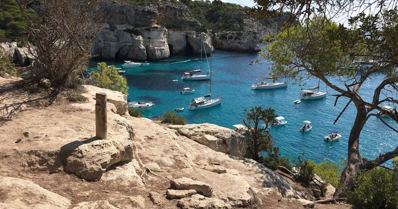 Een markeringspaal van het Camí de Cavalls op de rotsen boven Cala Macarella met boten in de turquoise baai op Menorca