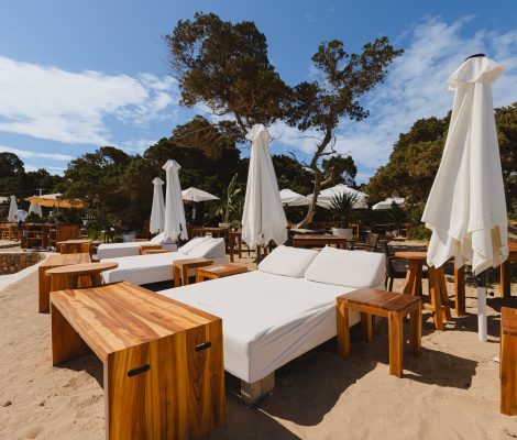 Balinese bedden en houten tafeltjes van Cala Bassa Beach Club op het zand tussen de bomen, met witte parasols