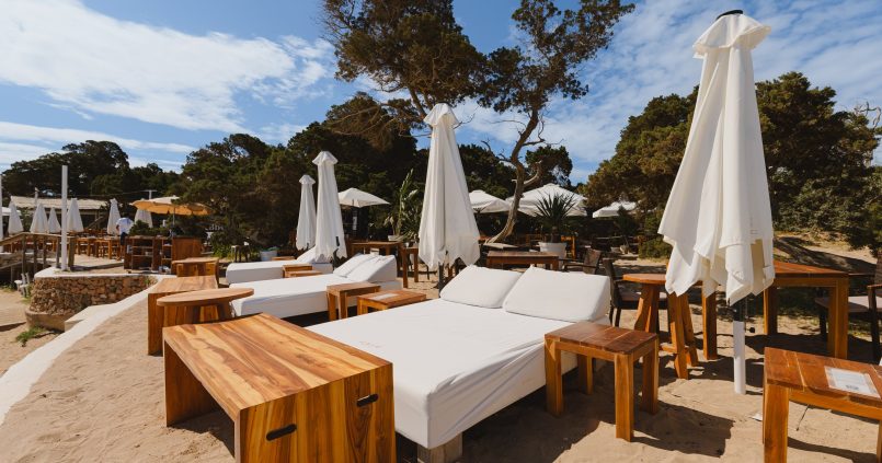 Balinese bedden en houten tafeltjes van Cala Bassa Beach Club op het zand tussen de bomen, met witte parasols