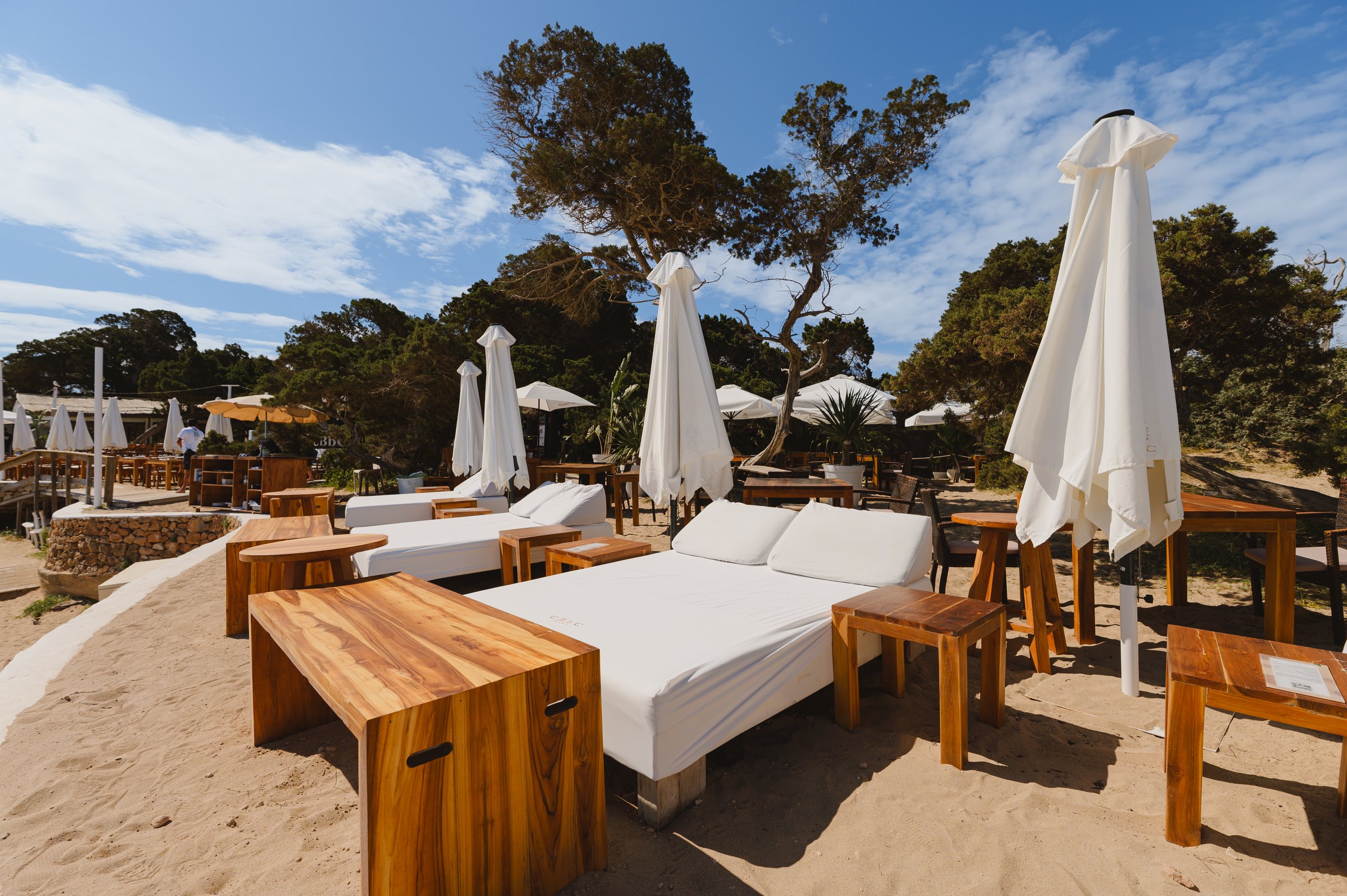 Cala Bassa Beach Club (CBbC) Ibiza: ligbedden, restaurants en prijzen 2026 1 Balinese bedden en houten tafeltjes van Cala Bassa Beach Club op het zand tussen de bomen, met witte parasols