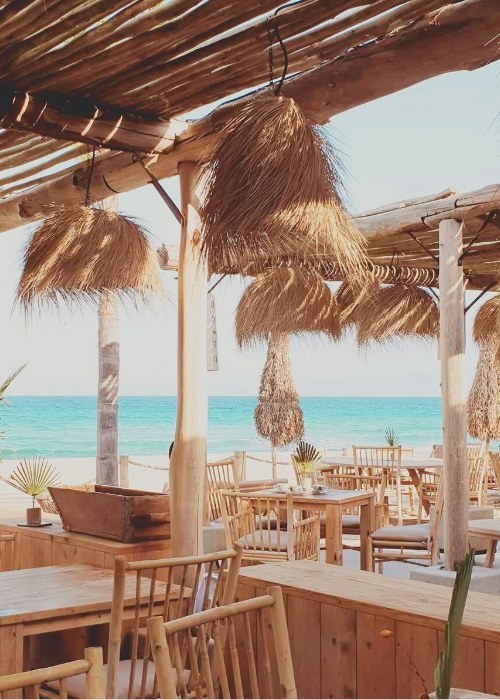Houten tafels en stoelen onder riet op het terras van Cucum Beach House