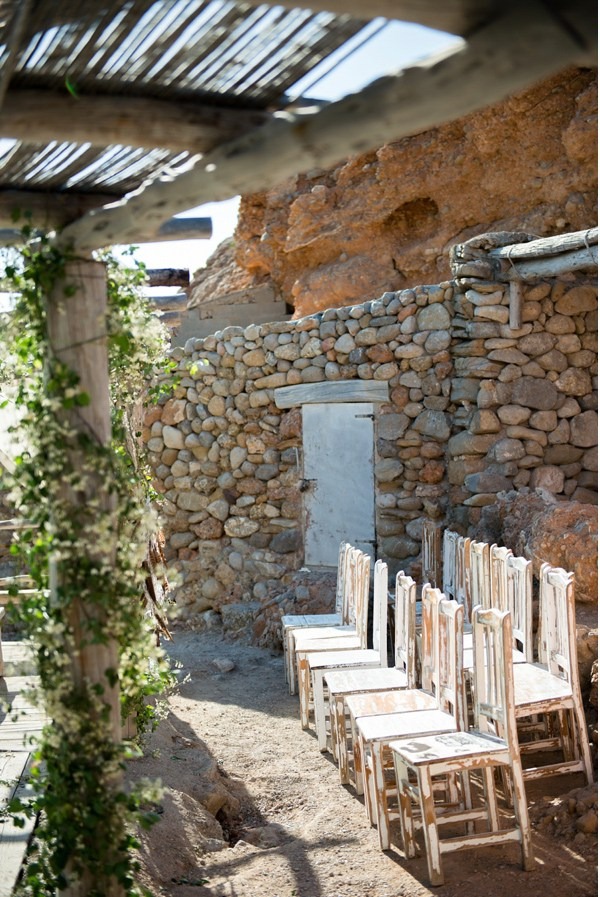 Rustieke houten stoelen tegen een stenen muur onder een rieten pergola bij Elements Ibiza