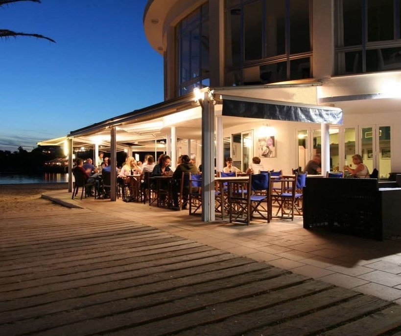 Het overdekte terras met tafels en blauwe stoelen van Es Bruc Santandría aan zee
